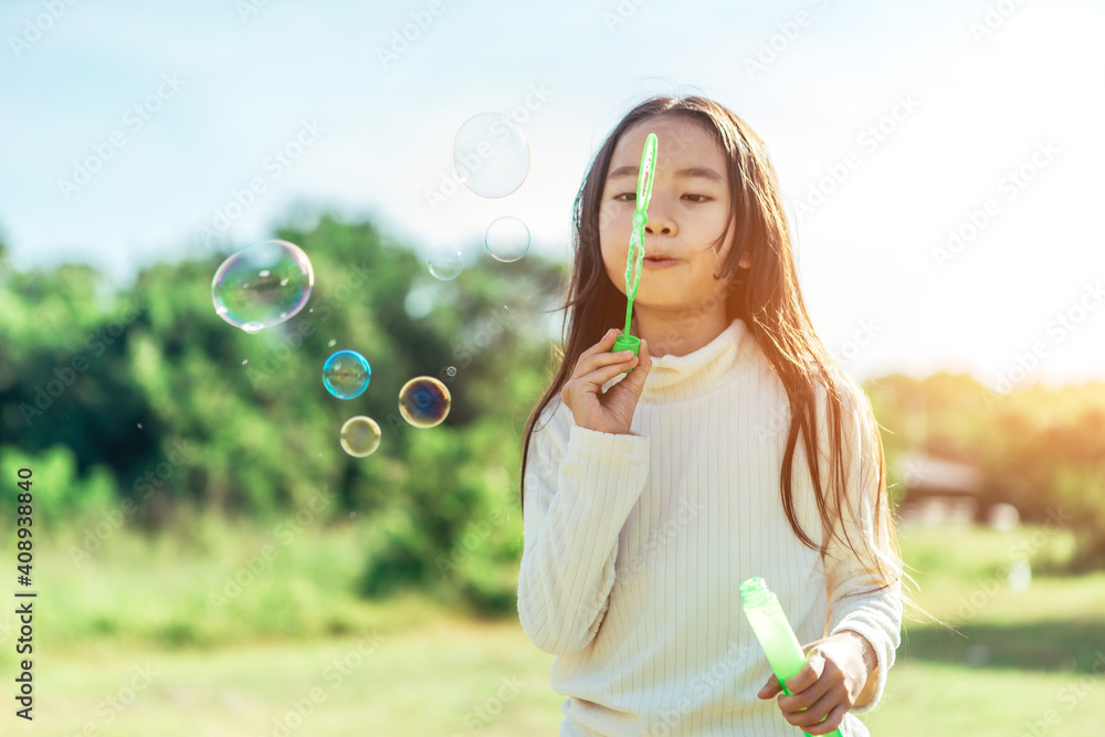 Portrait of funny lovely little Asian girl blowing soap bubbles in the park.