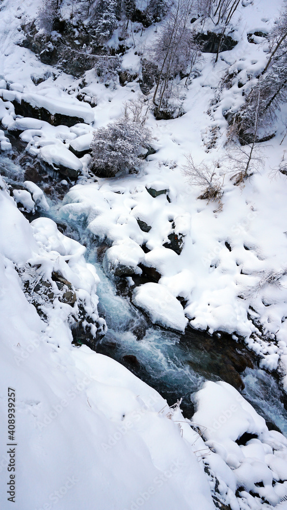 A clear mountain river runs through a snowy gorge. There is a metal bridge covered with snow. Tall spruce trees grow on the slopes of the mountains. There is steam from the river. Almarasan, Almaty A clear mountain river runs through a snowy gorge. There is a metal bridge covered with snow. Tall spruce trees grow on the slopes of the mountains. There is steam from the river. Almarasan, Almaty