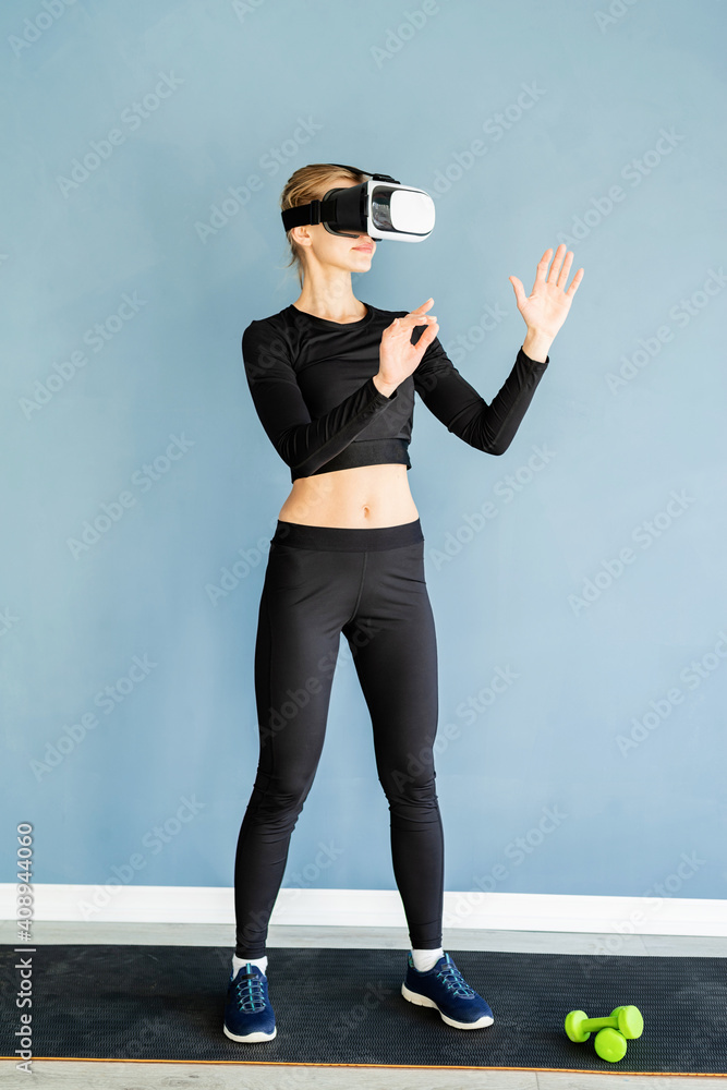 Young blonde woman in sport clothes wearing virtual reality glasses standing at fitness mat using VR interactive menu Young blonde woman in sport clothes wearing virtual reality glasses standing at fitness mat using VR interactive menu