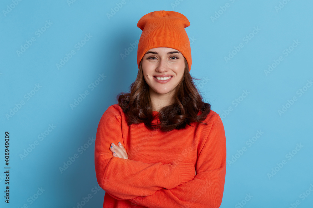 Beautiful young girl posing to camera with folded arms and charming happy smile, wearing orange casual sweater and cap, lady with dark hair against blue wall, expressing positive emotions. Beautiful young girl posing to camera with folded arms and charming happy smile, wearing orange casual sweater and cap, lady with dark hair against blue wall, expressing positive emotions.