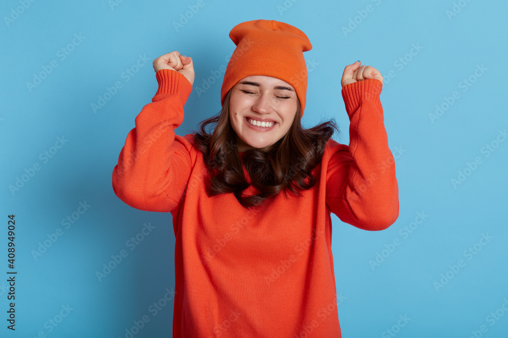 Happy lady dresses orange jumper and cap, keeps eyes closed and making hooray gesture, raising hands, feels very happy, celebrating success, posing against blue wall. Happy lady dresses orange jumper and cap, keeps eyes closed and making hooray gesture, raising hands, feels very happy, celebrating success, posing against blue wall.