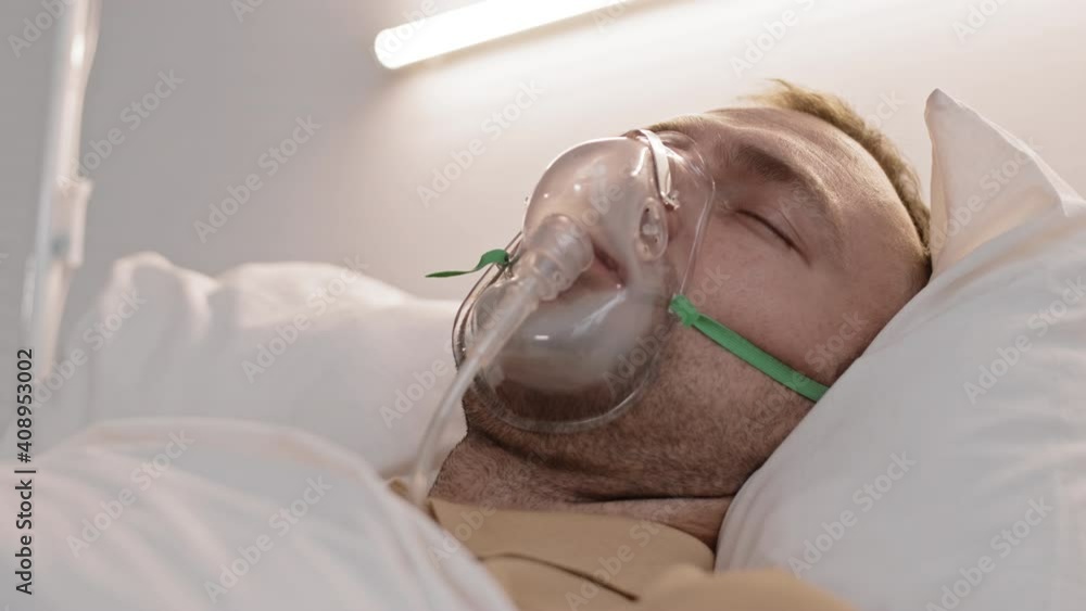 Low angle close-up of Caucasian man with oxygen mask on lying in bed with white linen, sleeping disturbingly, moving head