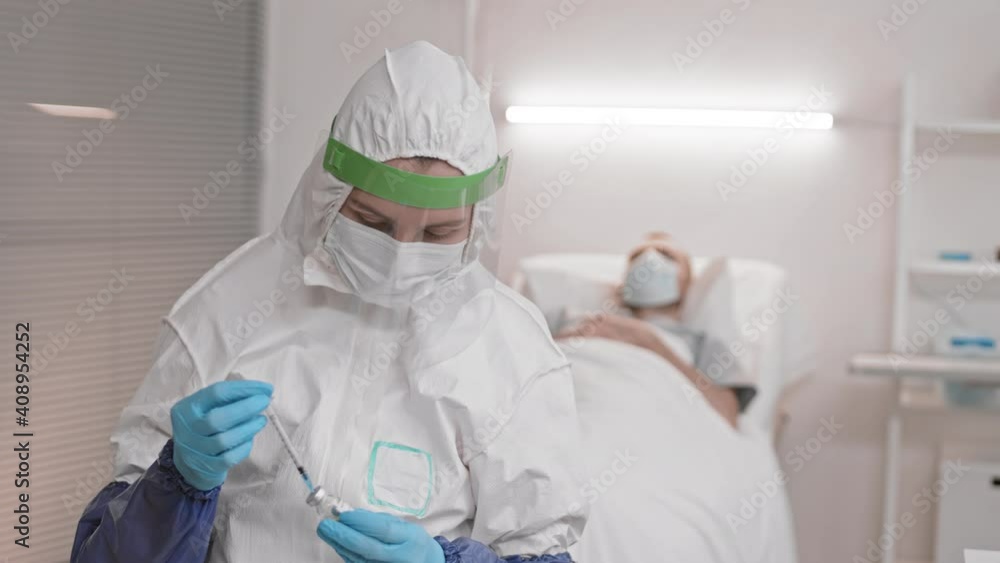 Waist up of young medical worker wearing full coverage hazmat suit and face shield filling in syringe with fluid from ampoule, standing in hospital ward of patient, then doctor looking up on camera Waist up of young medical worker wearing full coverage hazmat suit and face shield filling in syringe with fluid from ampoule, standing in hospital ward of patient, then doctor looking up on camera