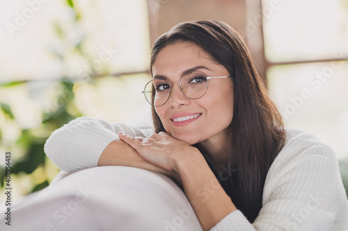 Photo of cheerful girl sit sofa rest head arms beaming smile wear eyeglasses white sweater in living room home indoors