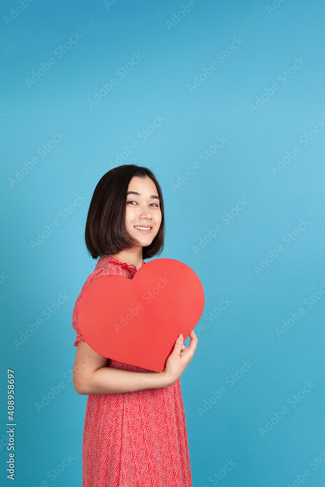 joyful, cheerful young asian woman in red dress holding big red paper heart like fan isolated on blue background. joyful, cheerful young asian woman in red dress holding big red paper heart like fan isolated on blue background.