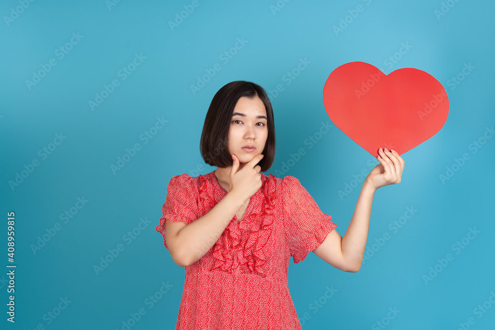 close-up thinking, decision-making young Asian woman in a red dress holds a large red paper heart in her hands and touches her chin with her hand , isolated on a blue background.