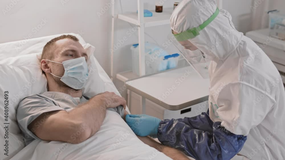 High angle of medical worker wearing full coverage protective suit giving injection in arm of hazardous Caucasian male patient lying in hospital bed. Man in face mask squinting eyes High angle of medical worker wearing full coverage protective suit giving injection in arm of hazardous Caucasian male patient lying in hospital bed. Man in face mask squinting eyes