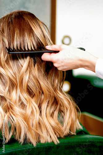 Back view of hairdresser combing wavy hair of a young blonde woman in a beauty salon