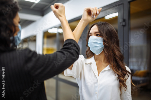 Safe greeting of two businesswomen wearing masks during coronavirus pandemic. Females cross their elbows to greet each other at an office corridor. Covid-19 spread prevention, new normal concept.