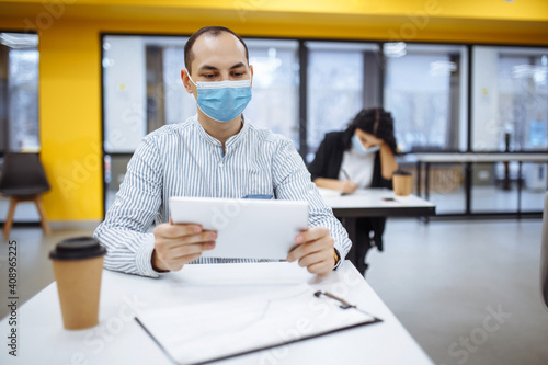 Young businessman working hard during pandemic of covid-19 at an office. Male professional sitting at a desk wearing medical mask, checking business trends. Health care and new normal concept.