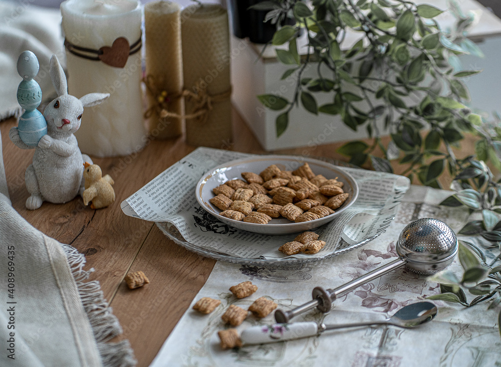 Easter composition. Scattered on a plate of sweets of square shape. against the background of candles, teaspoons of tea and Easter figurines with hare and eggs. Easter mood. Breakfast