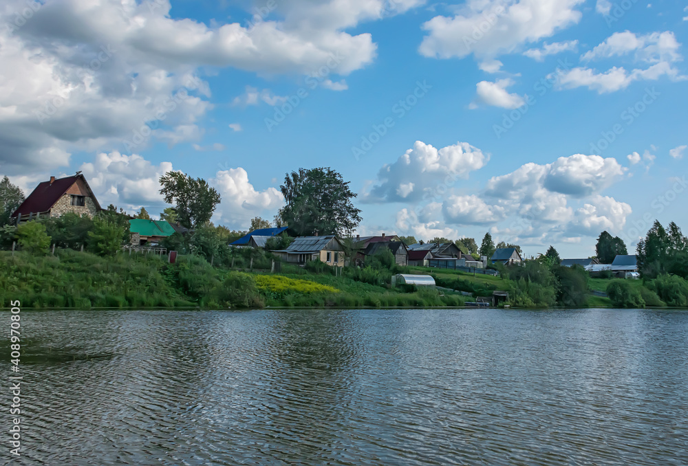 Obraz premium Village by the lake on a summer sunny day. By the lake, there are picturesque wooden houses with vegetable gardens and greenhouses, a blue sky with white cumulus clouds 