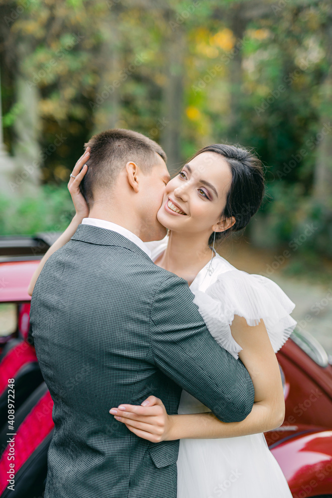 Portrait of happy newly wife and husband hugging outdoors and smiling, on background red recto car in tropical garden. Sincere feelings of two young people. Concept of true immortal love. Portrait of happy newly wife and husband hugging outdoors and smiling, on background red recto car in tropical garden. Sincere feelings of two young people. Concept of true immortal love.