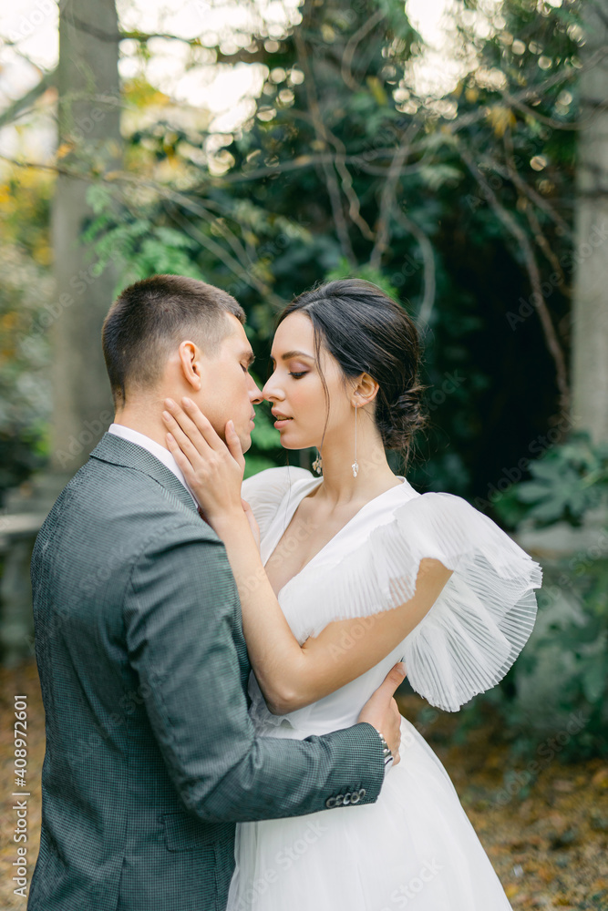 attractive bride in a delicate fashionable wedding dress kisses the groom in a gray suit near stone columns in a tropical garden on a warm summer day.