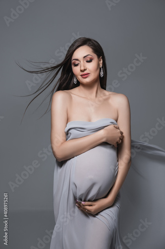 beautiful tender pregnant brunette woman in flying transparent grey fabric stands in studio