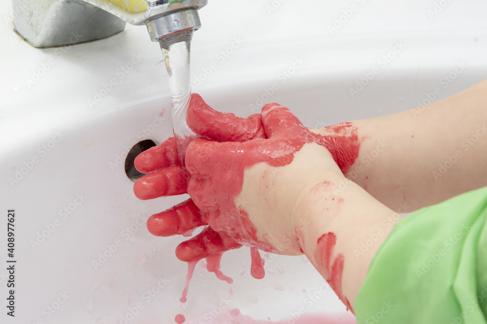 Fototapeta premium The girl washes her hands of red paint in the washbasin. Concept: children's educational games with paints, personal hygiene.