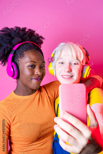 Two young multiethnic friends wearing on pink background taking selfie - Isolated diverse female streaming live using smartphone