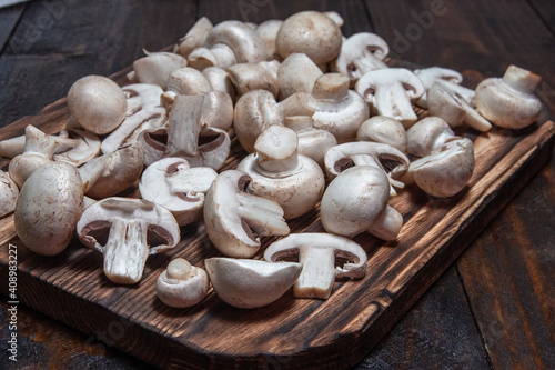 A pile of sliced and whole champignons lie on a burnt cutting board against a dark wooden background