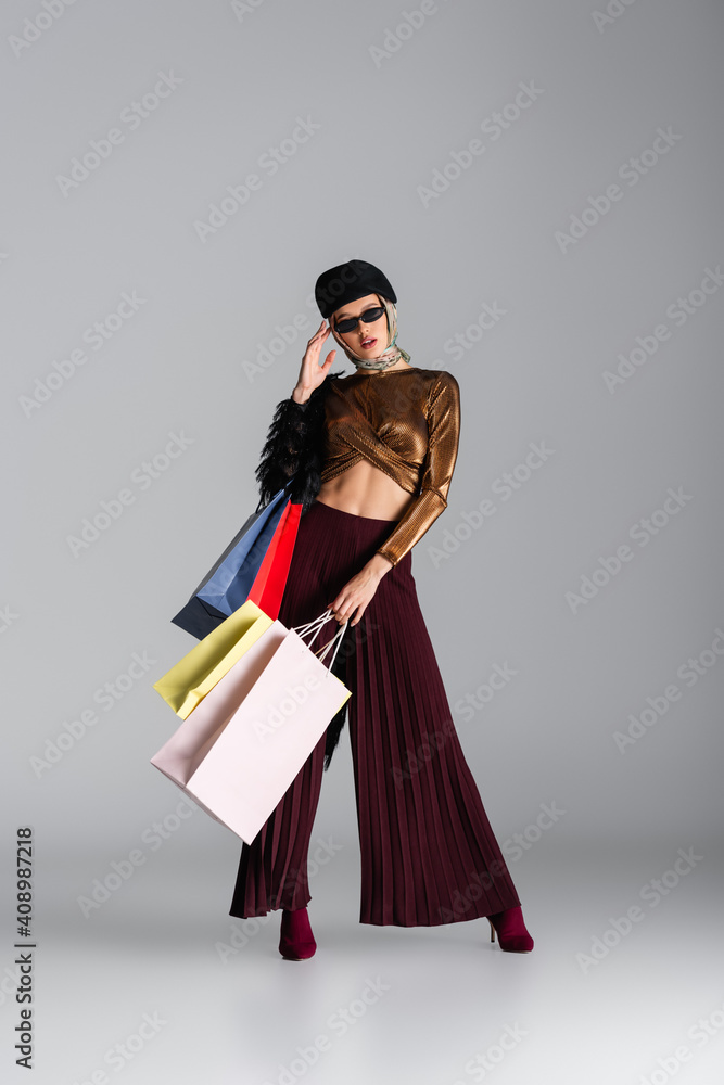 full length of fashionable young woman adjusting sunglasses and holding shopping bags while posing on grey