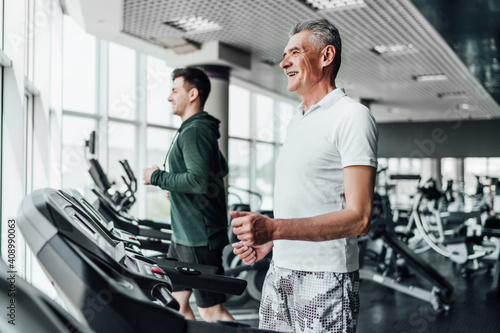 Two men on a treadmill in the gym, in focus a gray-haired, smiling senior man. Sports, training, health. Sports Hall