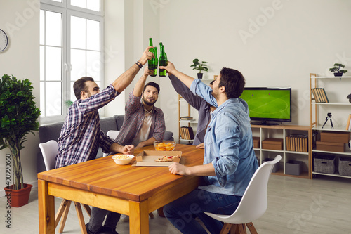 Cheers! Friends sitting at table at home and clinking beer bottles. Group of happy young men eating pizza, enjoying drinks, watching football match on TV, having fun and enjoying good time together