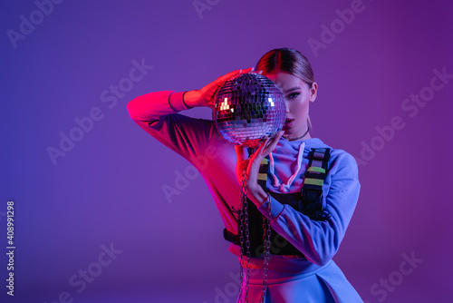 young and stylish woman covering face with shiny disco ball on purple