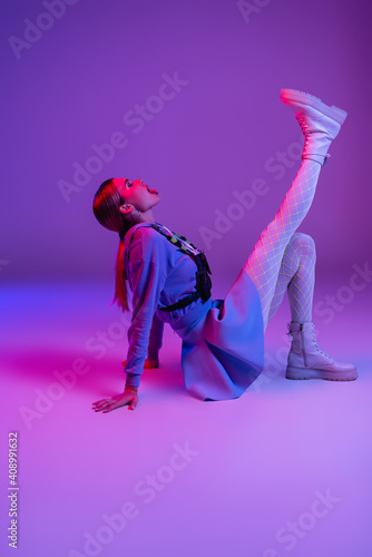 full length of stylish woman in trendy outfit sitting and sticking out tongue on purple