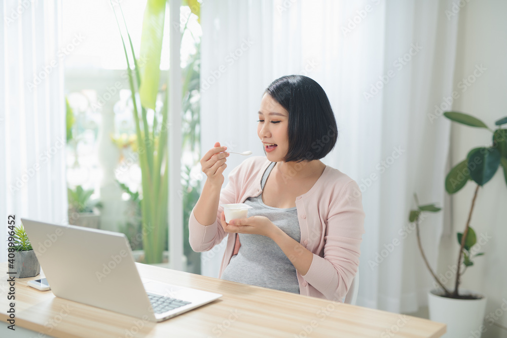 Beautiful pregnant business woman is eating yogurt and smiling while sitting at her working place at home Beautiful pregnant business woman is eating yogurt and smiling while sitting at her working place at home