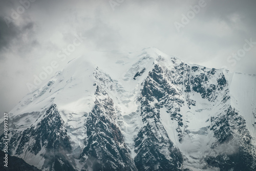 Atmospheric mountains landscape with big snowy mountain top with snow cornices in low clouds. Awesome minimal scenery with snow-white high pinnacle in overcast weather. Giant mountain wall in clouds.