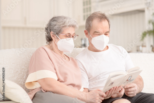 An elderly couple with medical masks are sitting on the couch at home and reading a book. Ways not to get bored during lockdown and develop while sitting at home. Quarantine during coronavirus pandemi