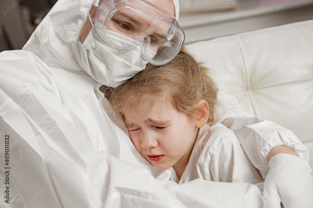 The doctor in a protective suit, mask, gloves and glasses calms down, hugs a little sick girl. children's tears. call the doctor at home. Pandemic.