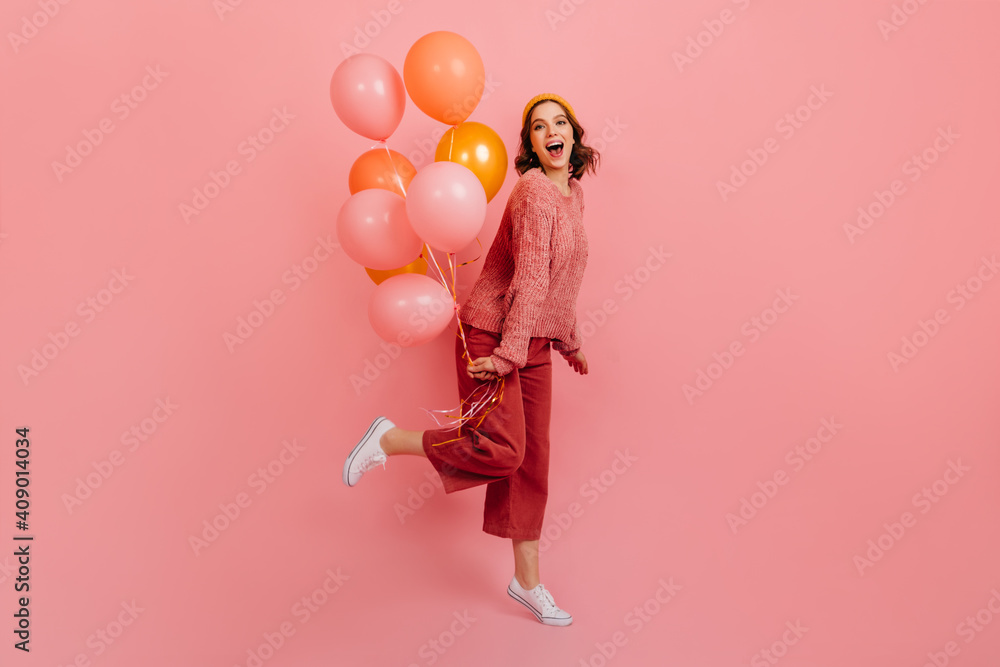 Full length view of joyful lady jumping with air balloons. Studio shot of laughing birthday girl posing on pink background.
