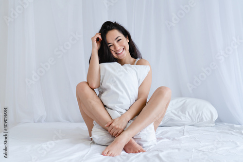 Attractive young caucasian woman sitting on the bed covering her body with a big white pillow