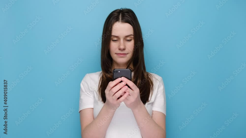 Portrait of cute lady enjoys distant communication, uses mobile phone, surfes fast unlimited internet, has pleasant smile, dressed in white t-shirt, makes shopping online, isolated on blue background Portrait of cute lady enjoys distant communication, uses mobile phone, surfes fast unlimited internet, has pleasant smile, dressed in white t-shirt, makes shopping online, isolated on blue background
