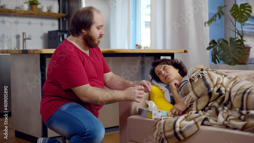 Adult man bringing glass of water for sick aged mother lying on couch