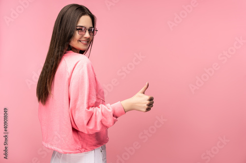 Photo of attractive young woman in glasses gives thumb up and smiles, wears casual pink t-shirt white pants isolated pink color background