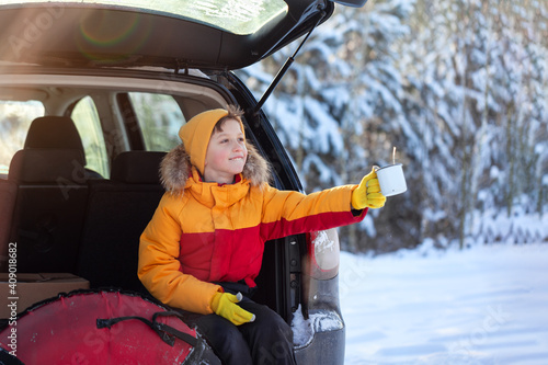 Adorable boy with hot tea or cocoa in his hands sitting in black car at winter day. Road trip,getaway, natural environment, staycation, travel, tourism at winter time. photo