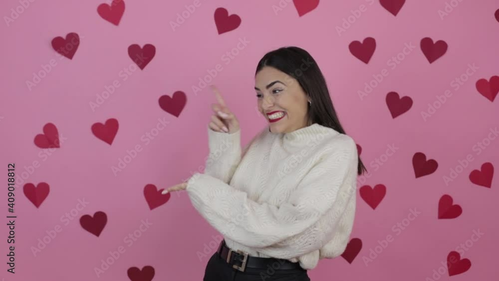 Young confident woman pointing aside on copyspace over isolated pink background with red hearts Young confident woman pointing aside on copyspace over isolated pink background with red hearts