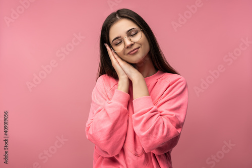 Photo of attractive young woman in glasses shows sleep, wears casual pink t-shirt white pants isolated pink color background © Ivan