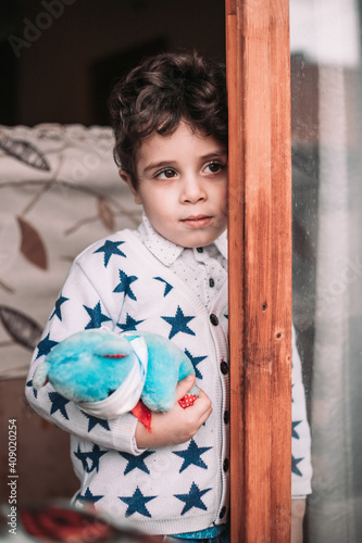 Multiracial boy holding a teddy bear with a mask on. Standing by the window