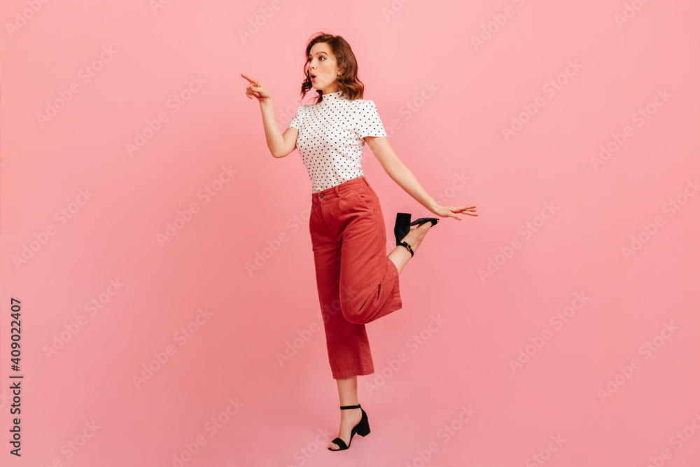 Full length view of shocked woman pointing with finger. Emotional lady in stylish pants standing on pink background. Full length view of shocked woman pointing with finger. Emotional lady in stylish pants standing on pink background.