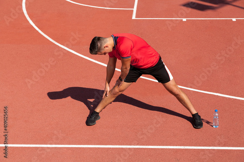 Handsome sportsman stretch his legs and preparing on red terrain, during sunny day, wearing red t shirt and black sneakers