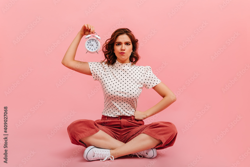 Dissatisfied girl showing time on pink background. Studio shot of pretty woman sitting on floor with clock. Dissatisfied girl showing time on pink background. Studio shot of pretty woman sitting on floor with clock.