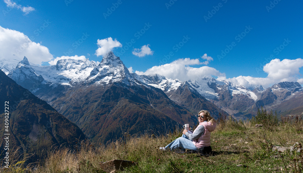 Fototapeta premium woman traveler drinks coffee with a view of the mountain landscape. A young tourist woman drinks a hot drink from a cup and enjoys the scenery in the mountains. Trekking concept.