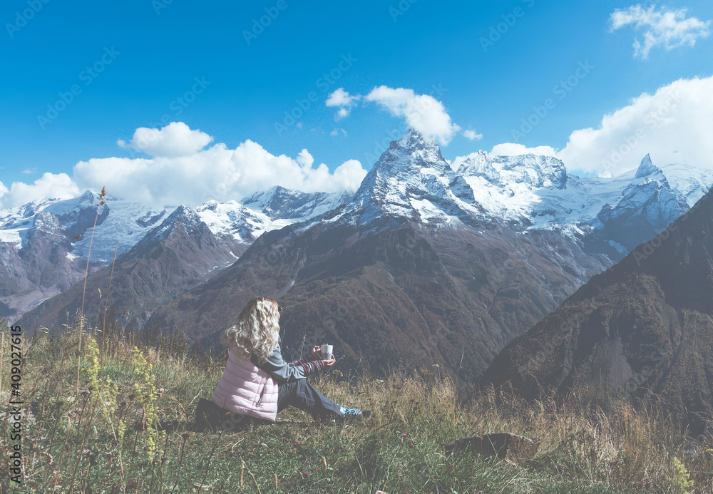 Fototapeta premium woman traveler drinks coffee with a view of the mountain landscape. A young tourist woman drinks a hot drink from a cup and enjoys the scenery in the mountains. Trekking concept.
