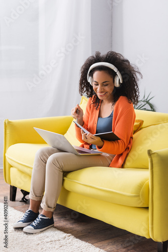cheerful african american freelancer in wireless headphones using laptop in living room