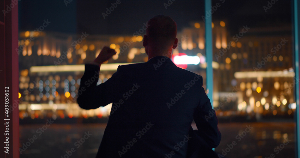 Back view of businessman celebrating success standing near panoramic window with night cityscape - obrazy, fototapety, plakaty 