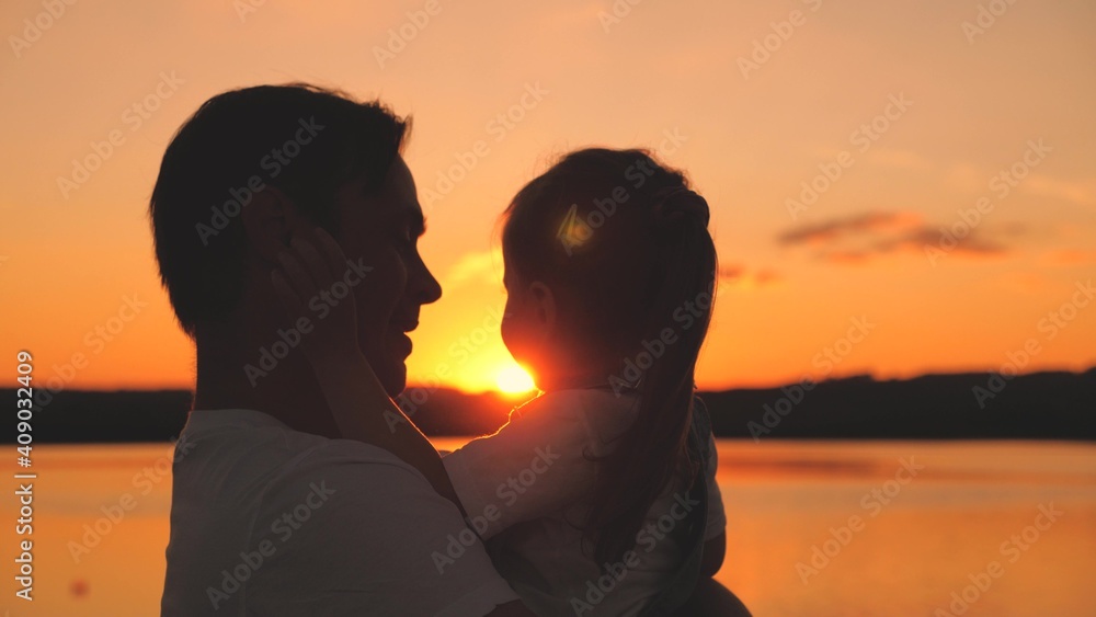 Silhouette of a dad hugs his little daughter at sunset in the sky and smiles. Happy Dad's Day. The girl sits in her father's arms at dawn and laughs. Family life with a child. Raise kid. Childhood