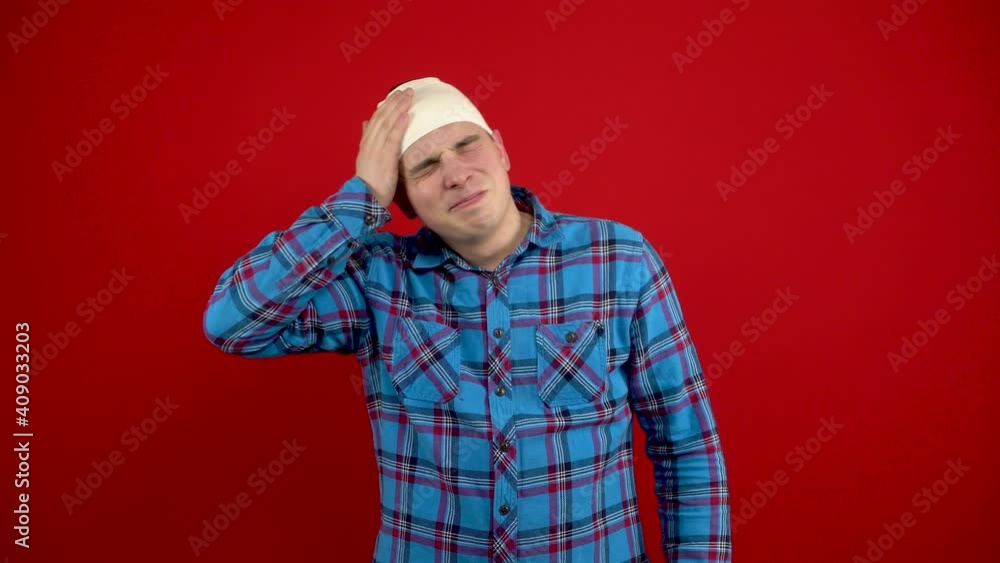 The young man has a bandage on his head, he is holding it with his hands because of the pain. Shooting on a red background. The young man has a bandage on his head, he is holding it with his hands because of the pain. Shooting on a red background.