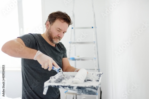 Portrait of a handsome mature man making repairs in the apartment. The person paints the wall white with roller. Do it yourself. DIY